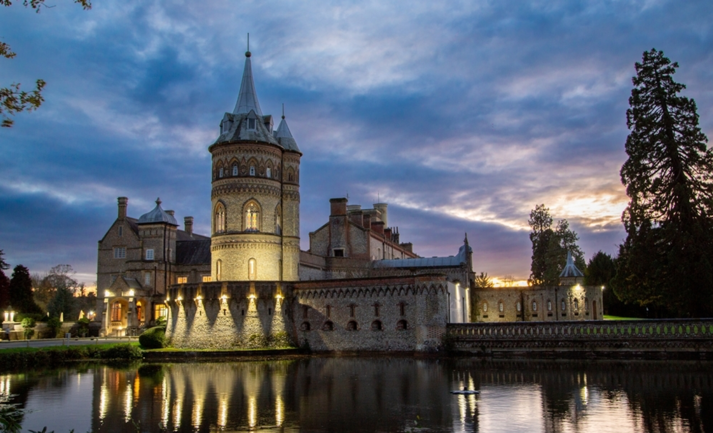 De Vere Horsley Estate lit up at dusk, its historic tower reflected in the lake — the Surrey venue for the Heal Money Trauma Retreat